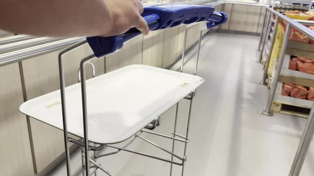Close view of a person pushing a small shopping cart with a white tray in a bright self service store or cafeteria aisle, showing everyday shopping, convenience, and modern retail.
