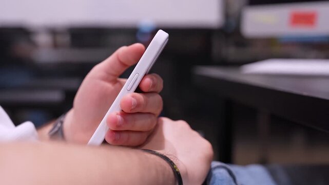 Person holding white smartphone and using touch screen to browse internet in modern office space.
