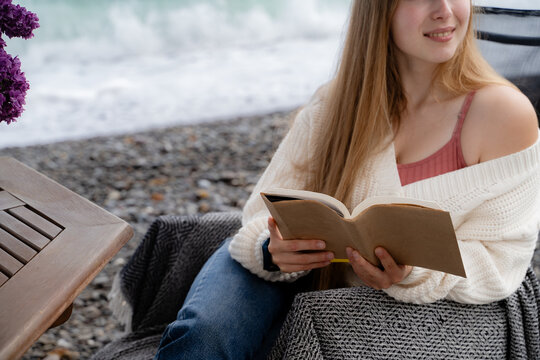 A relaxed smiling woman is reading a book on a pebbled beach near the sea during a peaceful outdoor break.
