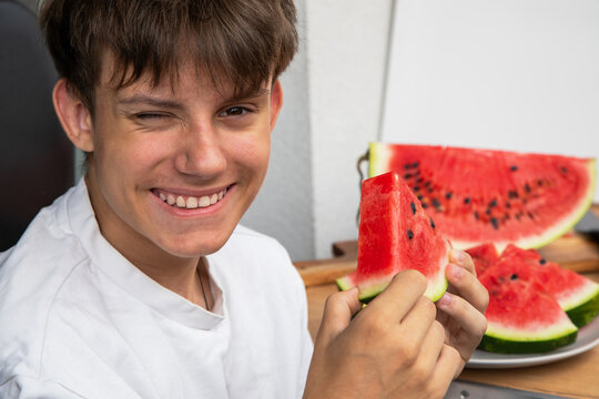 Happy beautiful teenager enjoys sweet juicy watermelon slice. A cheerful boy winking and smiling while holding a fresh slice of ripe red watermelon. Perfect summer snack and healthy lifestyle concept 