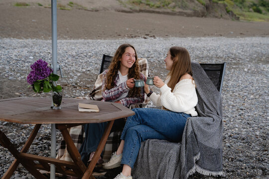Two cheerful women are relaxing at a seaside table, enjoying a friendly tea break and the coastal lifestyle. Happy girls are chatting and enjoying each other's company on the beach.