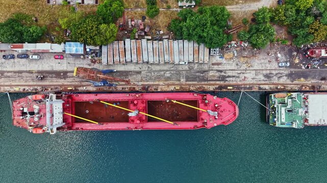 Multiple ships are docked at a busy port with loading and unloading of cargo taking place. Several workers and equipment are visible along the shore near the water.