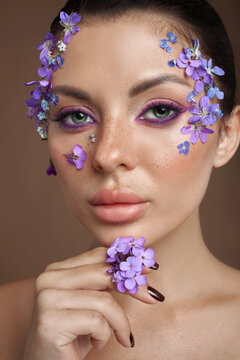 A model shows off a beauty look using flowers on her face and hand. She has purple eye makeup and flowers placed carefully around her features. The setting is a studio