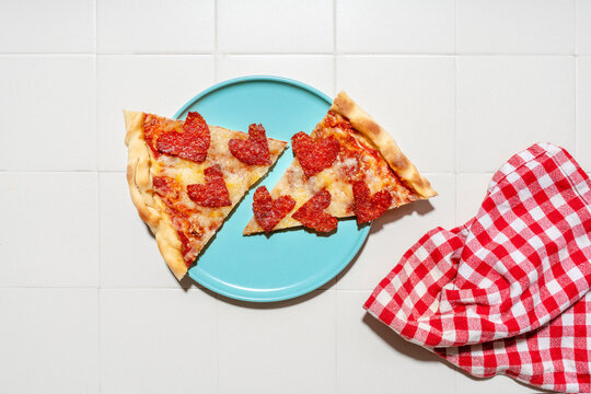 Pizza with pepperoni in heart shapes on blue plate, white tiles and white and red checkered napkin