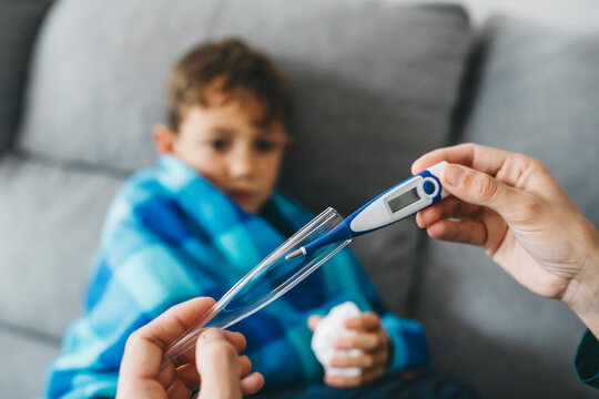 Mother's hand taking digital thermometer while sich son waiting on couch in the background