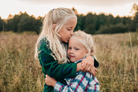 A blonde girl hugs and kisses her sister in a summer meadow