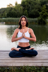 Fototapeta premium A young Hispanic woman practises a seated lotus meditation on the grass beside the water in the evening light, a moment of self care and active summer living by the lakeside.