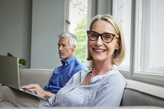 Portrait of smiling mature woman on couch with man in background using tablet