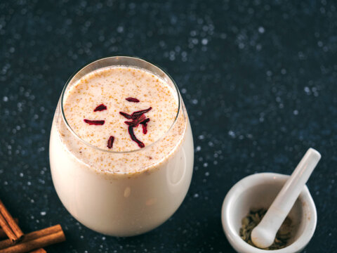 Traditional spiced milk beverage Sardai with saffron threads on top. Served in a glass on a dark background with mortar and cinnamon sticks. Copy space, perfect for Holi celebration marketing