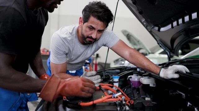 Two auto technicians collaborate over an open car hood, inspecting high voltage wiring and components during electric vehicle maintenance in a garage.