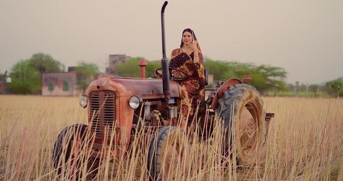 Indian young adult woman sitting on tractor learn driving steering wheel look camera at farm rural area. Happy female wear saree staring cam do casual pose enjoy desi life day village ground place