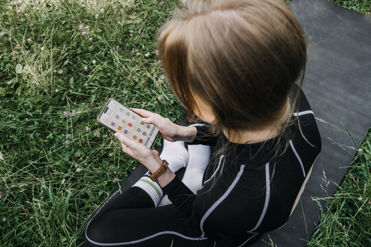Woman sits on mat in grass holding smartphone with screen showing wellness habits app. Mobile mental health trackers, daily self care routines, digital habit building, somatic wellness therapy.