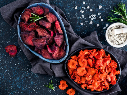 Baked beet and carrot slices with coarse sea salt scented rosemary. Vegan diet food idea and recipe. Healthy homemade beetroot and carrot chips in plate and ingredients on dark blue background. Copy
