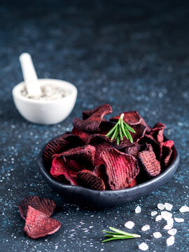 Homemade crispy vegan beet slices snack in a black bowl. Dark blue textured background with coarse sea salt, rosemary sprigs and copy space. Perfect for healthy diet and organic food marketing