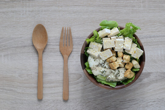 Top view of healthy salad bowl with tempeh, croutons, and green lettuce, served with wooden spoon and fork on a rustic table background.