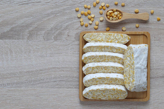 Fresh raw tempeh slices on a wooden tray with a small spoon of soybeans, healthy plant-based fermented protein food ingredient, top view.