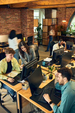 Employees working at computers while blurred coworkers cross busy open plan office. Concept of office multitasking, hybrid work environment and modern business operations.