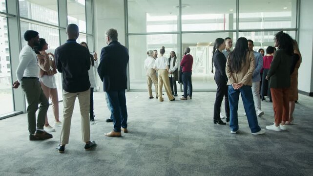 Diverse business professionals engage in conversations, fostering connections and networking in a spacious, glass-walled venue. They stand in small groups, sharing ideas and building relationships.
