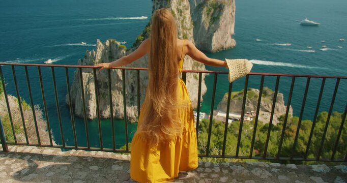 Woman in sundress admiring Faraglioni cliffs view from hilltop terrace on Capri Island in summer sunny day