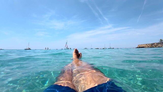 First person point of view of a man relaxing and floating in the crystal clear turquoise water of tabarca island, alicante, spain, with sailboats in the background on a sunny day