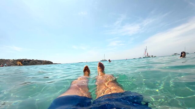 First person perspective of a man relaxing and floating in the beautiful turquoise waters of tabarca island, spain, during a sunny summer vacation with sailboats in the background