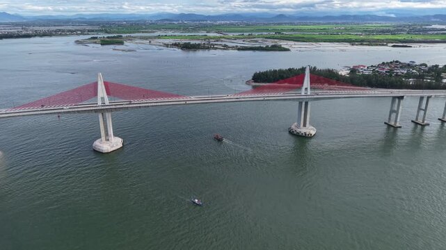 Close aerial shot circles one of the Thuan An Bridge main towers, showing the radiating red cable stays supporting the bridge deck over the open water.