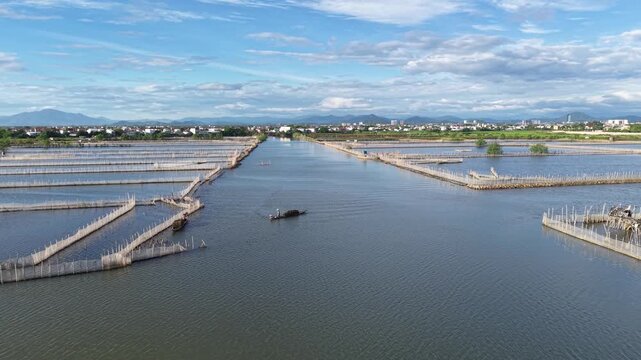 Aerial view shows a worker in a small boat tending aquaculture ponds on the lagoon, the pond borders and nets visible from above.