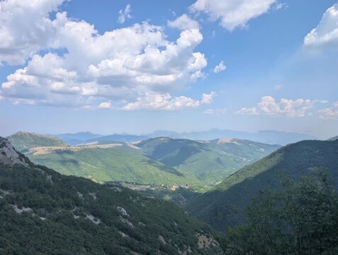 Vista ampia su pendii verdi e vallate profonde illuminate da luce naturale, con montagne che sfumano all&rsquo;orizzonte sotto un cielo parzialmente nuvoloso