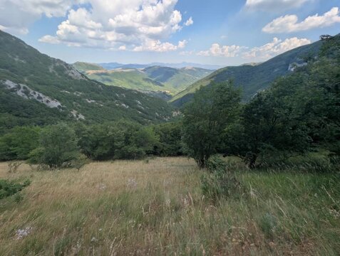 Vista ampia su pendii verdi e vallate profonde illuminate da luce naturale, con montagne che sfumano all&rsquo;orizzonte sotto un cielo parzialmente nuvoloso