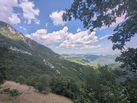 Vista ampia su pendii verdi e vallate profonde illuminate da luce naturale, con montagne che sfumano all&rsquo;orizzonte sotto un cielo parzialmente nuvoloso