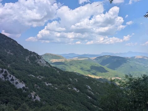Vista ampia su pendii verdi e vallate profonde illuminate da luce naturale, con montagne che sfumano all&rsquo;orizzonte sotto un cielo parzialmente nuvoloso