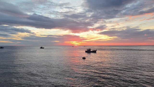 Aerial view captures several fishing boats heading out to sea at dawn, their wakes creating patterns on the calm water surface under a colorful sunrise sky.