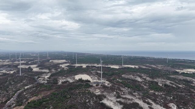 Aerial view of wind turbines rising above scattered coastal settlements and dry scrubland, showing the coexistence of wind energy infrastructure and rural communities.