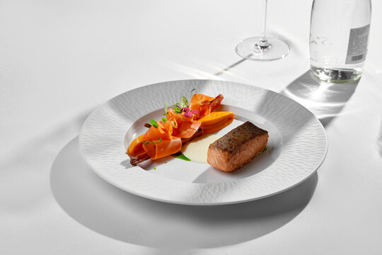 Seared salmon with carrot ribbons on white plate. Sunny minimalist fine dining table setting.