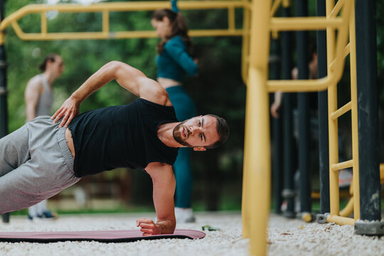 A fit man holds a side plank on a mat at an outdoor exercise area. The scene shows an outdoor workout with people training in the background.