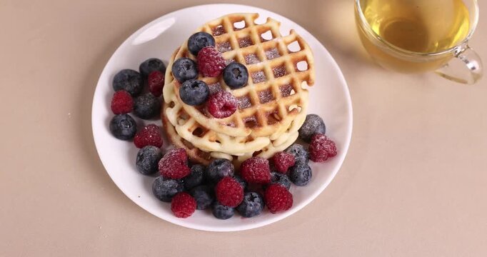 Stack of golden waffles with mixed berries on a white plate, cup of tea nearby, colorful breakfast display, appetizing food presentation, studio scene
