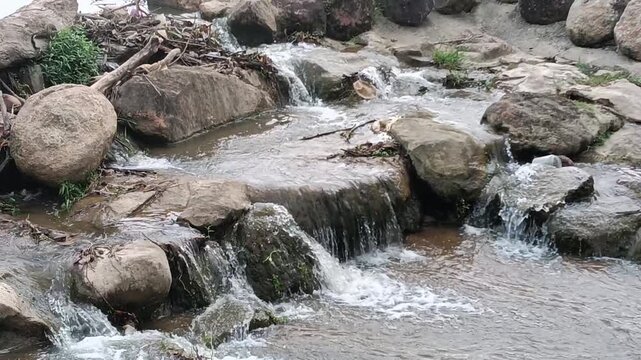 A small rocky stream with cascading water flowing over smooth boulders in a natural outdoor setting. The scene features clear running water, weathered rocks, patches of green vegetation, driftwood, an