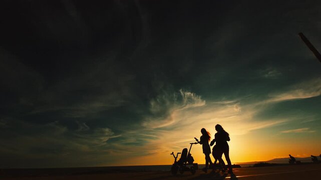 Group of women walking along a beachside promenade at sunset, with silhouettes against a vibrant sky and a stroller visible in the foreground
