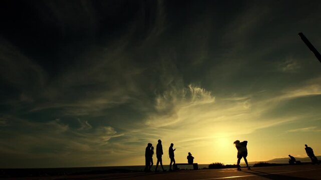 Silhouetted figures skateboarding and walking along a coastal pathway at sunset, with vibrant clouds and a glowing horizon creating a picturesque scene