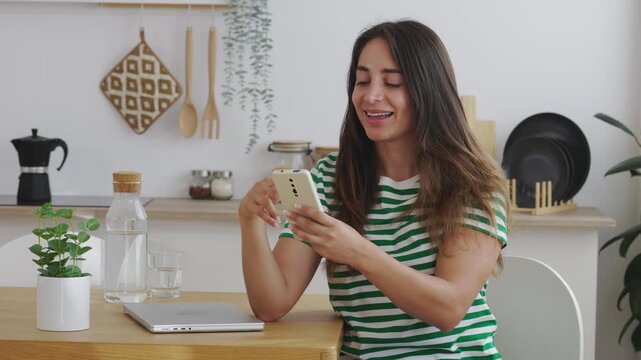 Beautiful young Latin American woman with long hair sitting at her kitchen table, smiling and laughing while scrolling through her social media feed on a modern smartphone, enjoying her leisure time