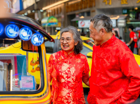 Couple in red outfits smiling by a colorful tuk-tuk at a busy street market in Bangkok during the daytime