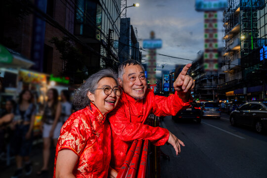Elderly couple in traditional clothing enjoy evening street festival and point at something interesting in busy city