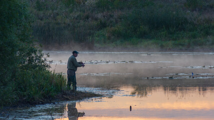 A fisherman stands on a misty riverbank during a serene sunrise, casting his fishing line into the calm, reflecting water © Александр Арендарь