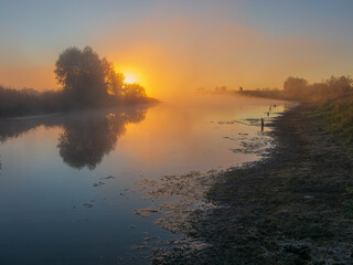 Naklejka premium A golden sunrise illuminates a misty river, casting warm light and reflections on the calm water and its grassy banks