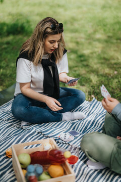 Young woman sitting cross-legged on a striped picnic blanket holding playing cards. A casual outdoor picnic scene with friends playing cards on the grass.