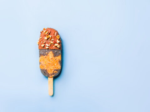 Healthy homemade plant-based chia seeds popsicle with raw carrot cake layer and chocolate coating with nuts. Minimalist flat lay on blue background. Chia popsicle with chocolate, copy space