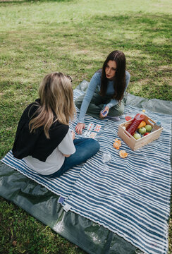 Two women sit on a striped picnic blanket playing cards and sharing fruit. A relaxed outdoor picnic scene of friends socializing in a grassy park.