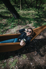 Obraz premium A young woman lies in a hammock outdoors wearing sunglasses and offering small wildflowers toward the camera. She relaxes in a shaded forest setting, enjoying a peaceful moment in nature.