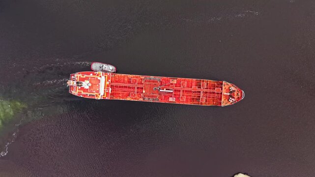 A large cargo ship is being guided by a tugboat across dark water. The scene shows activity in a shipping route. Other vessels may be nearby as it navigates carefully.