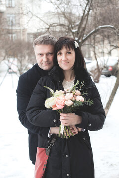 A couple is standing on a snowy street. The woman holds a bouquet of flowers and smiles. The man stands beside her in formal attire.